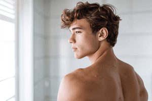 Young man with curly hair showing his back, highlighting skin texture in a bright bathroom.