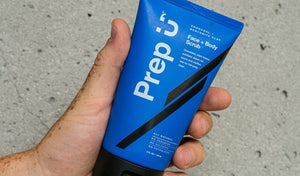 Hand holding a blue tube of Prep U charcoal face and body scrub against a textured background.