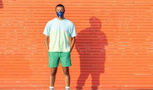 Young man standing confidently against an orange background, wearing a colorful shirt and mask.
