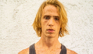 Close-up of a teenage boy with blonde hair and freckles against a textured wall.