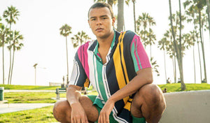 Young man in a colorful striped shirt squatting at a sunny park with palm trees.