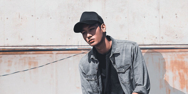 Young person in a cap and denim jacket posing against a rustic wall, radiating cool confidence.