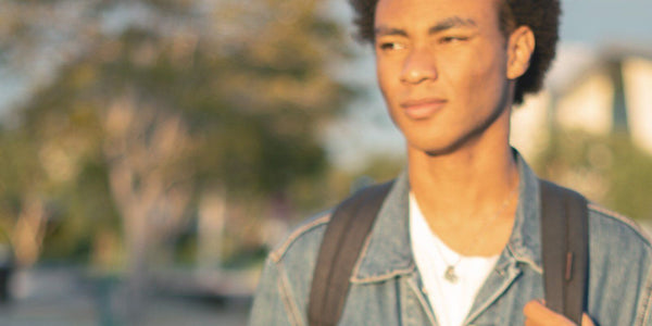 Young man with curly hair, wearing a denim jacket and backpack, enjoying a sunny day outdoors.