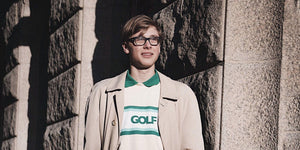 A young man wearing glasses and a GOLF shirt stands stylishly against a stone wall, showcasing modern fashion.