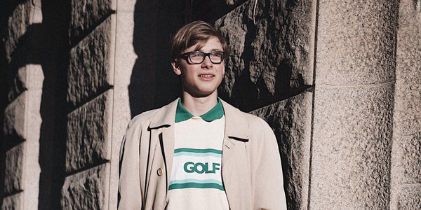 A young man wearing glasses and a GOLF shirt stands stylishly against a stone wall, showcasing modern fashion.