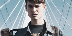 Teen boy with stylish haircut wearing a denim jacket, standing confidently under a modern bridge.