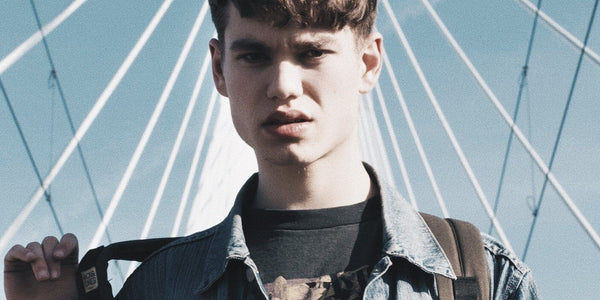 Teen boy with stylish haircut wearing a denim jacket, standing confidently under a modern bridge.