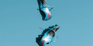 Dynamic shot of blue New Balance cleats flying against a clear blue sky, showcasing sporty style and performance.
