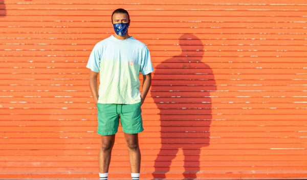 Young man standing confidently against an orange background, wearing a colorful shirt and mask.