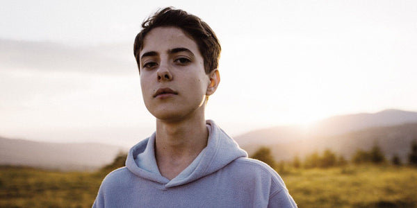 Teenage boy with a serious expression standing in a field during sunset, wearing a light hoodie.