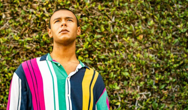 Stylish individual in a vibrant striped shirt standing against a green hedge, exuding confidence.