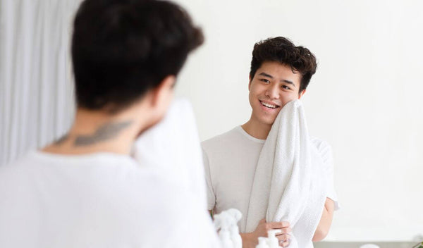 Teen boy smiling while drying his face with a towel in front of a mirror, showcasing a skincare routine.