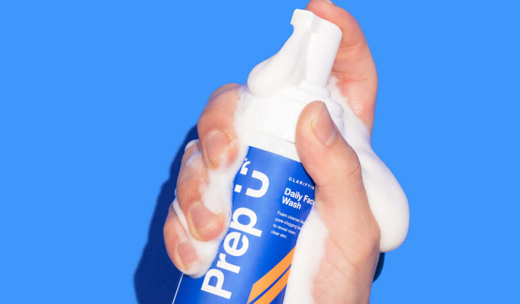 A hand dispensing fluffy foam from Prep U's Daily Face Wash bottle against a bright blue background.