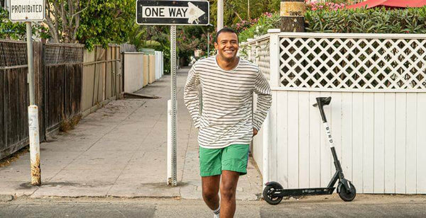 Young man smiling while walking along a street with a one-way sign and an electric scooter nearby.