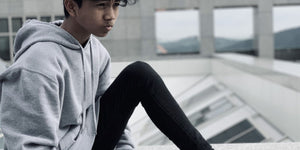 Teen boy in a gray hoodie sitting on a rooftop, looking contemplative against a city backdrop.