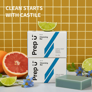 Two boxes of Prep U cleansing bars with a bar of soap in front, surrounded by citrus fruits and flowers on a tiled background.